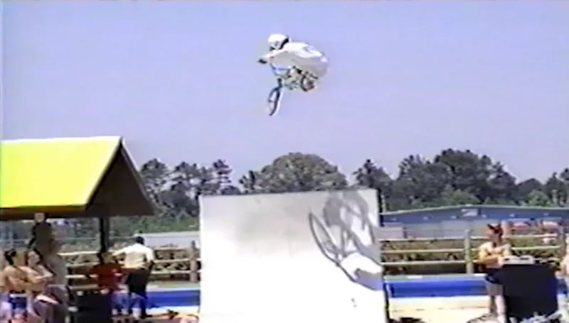 Kelly high above a vert ramp, tucked on the bike, the bike&rsquo;s shadow on the ramp face below and the crowd along the fence