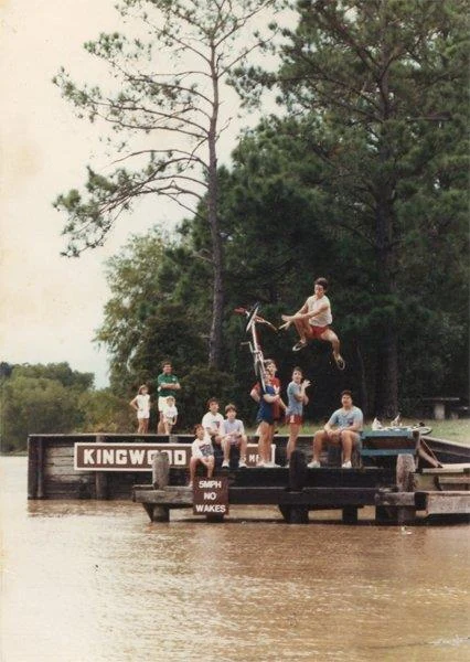 A teenage Kelly high in the air on a BMX bike, launched off a lakeside dock, with a group of kids watching from the dock below