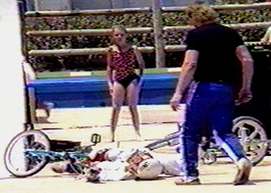Kelly lying on the concrete at the base of the ramp with the bike across his legs; a child and a man standing nearby