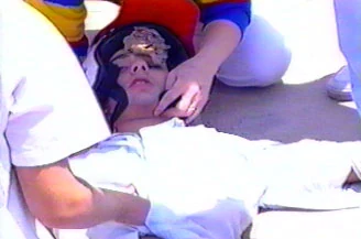 Kelly on his back on the concrete, helmet still on, a nurse's hands holding his head still