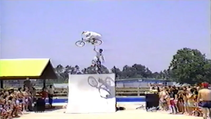 Kelly inverted high above the ramp while a teammate stands on the deck beneath him holding a bike overhead; the crowd along both sides of the ramp