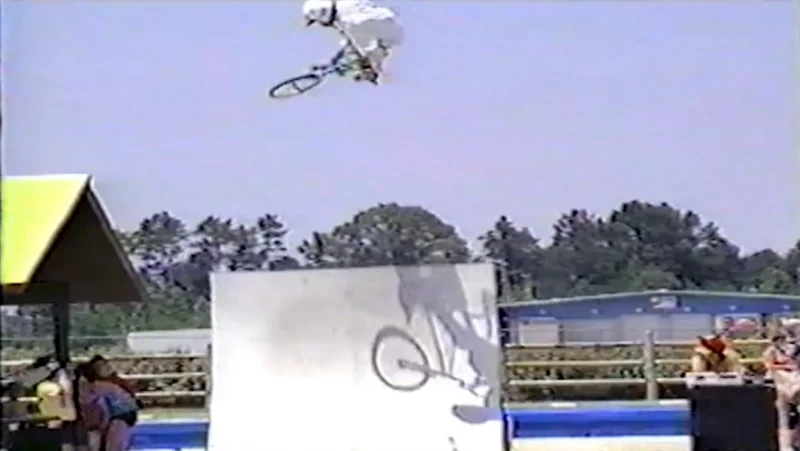 Kelly far above the vert ramp on the borrowed bike, the bike turned sideways at the top of the air, his shadow and the bike&rsquo;s on the ramp face below